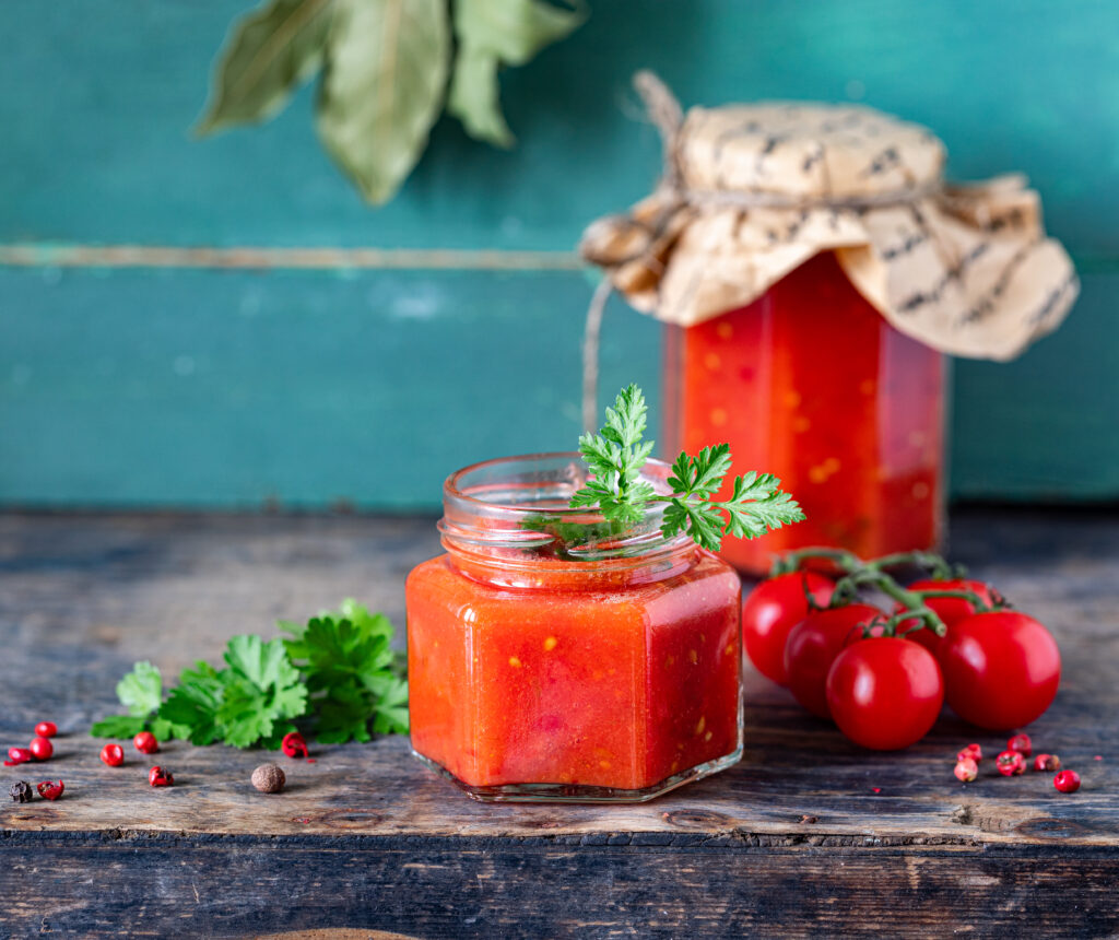 Homemade tomato ketchup made from ripe red tomatoes in glass jars with ingredients on an old wooden table